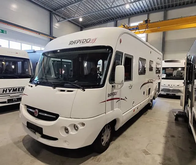 Hvit bobil av merke Rapido står innendørs sammen med flere andre bobiler i en salgsutstilling.