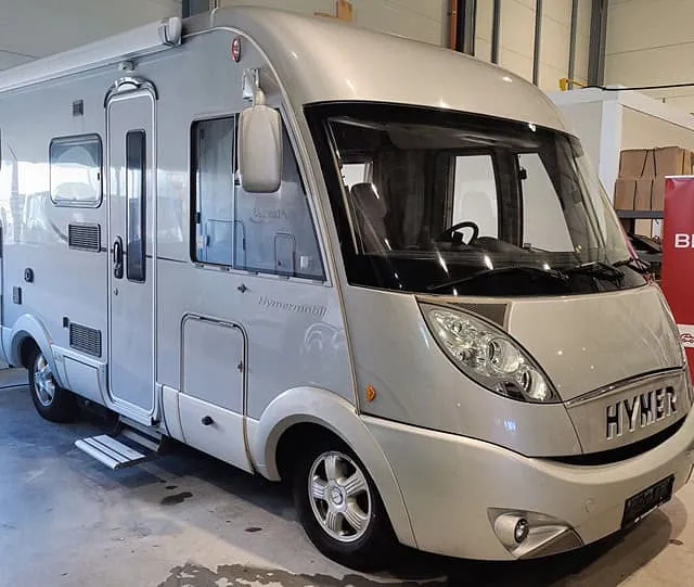 Kompakt sølvfarget Hymer-bobil parkert i innendørs salgsområde ved Brage Finans-banner.